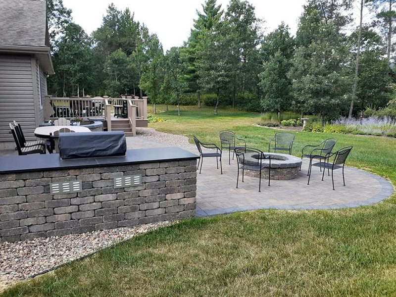 Paver patio with a stone fire pit seating area and built-in outdoor kitchen wall adjacent to a backyard deck in Nekoosa, Wisconsin